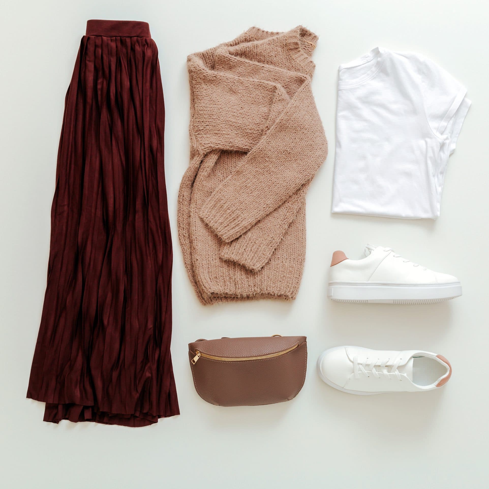 A complete outfit laid flat: burgundy skirt, camel sweater, white tee, white sneakers and a belt bag